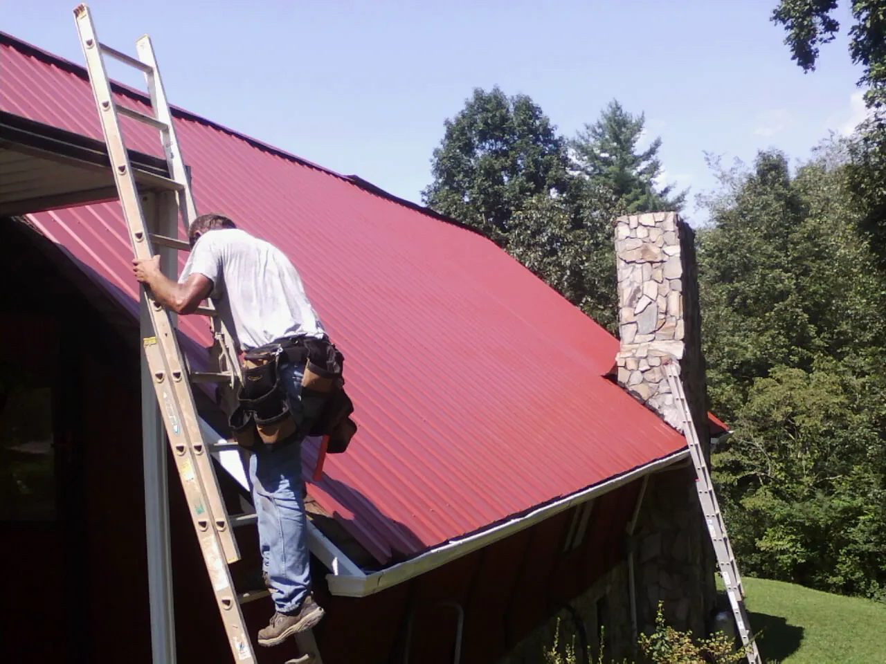 Roofer on a ladder beside a red metal roof and stone chimney — Soffit & Fascia Repair by Roofing Brothers in Palm Desert